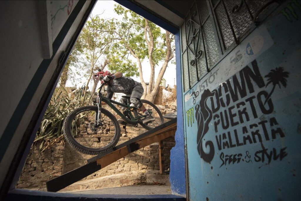 Atleta de downhill saliendo de una casa tradicional de Puerto Vallarta hacia una rampa de madera en el vibrante circuito urbano del centro.